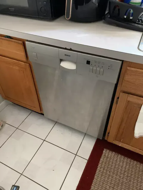 Stainless steel dishwasher under a kitchen counter, flanked by wood cabinets and tiled floor.