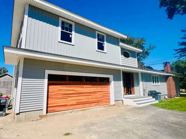 Front view of a two-story house with gray siding, a wooden garage door, and a small set of stairs leading to the front door. Clear blue sky in the background.