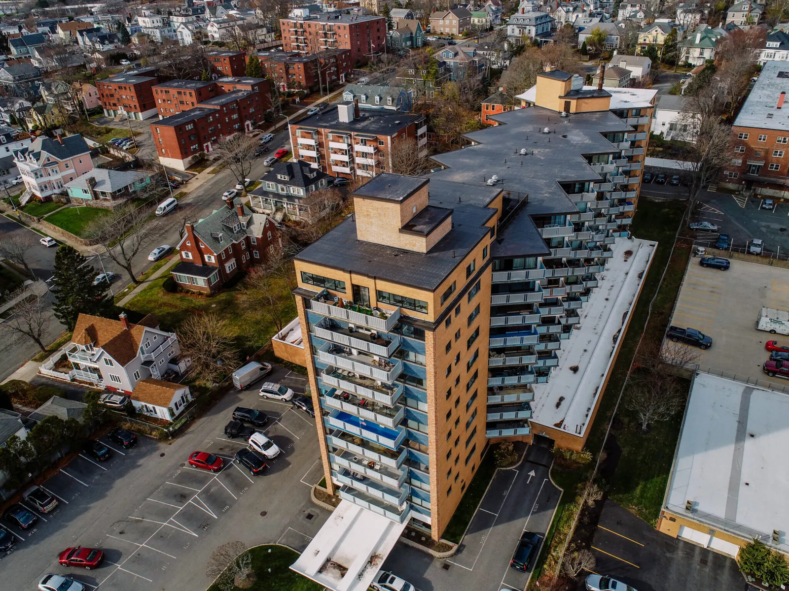Aerial view of a mid-rise apartment building with balconies, surrounded by parked cars, houses, and other residential buildings in a suburban neighborhood.