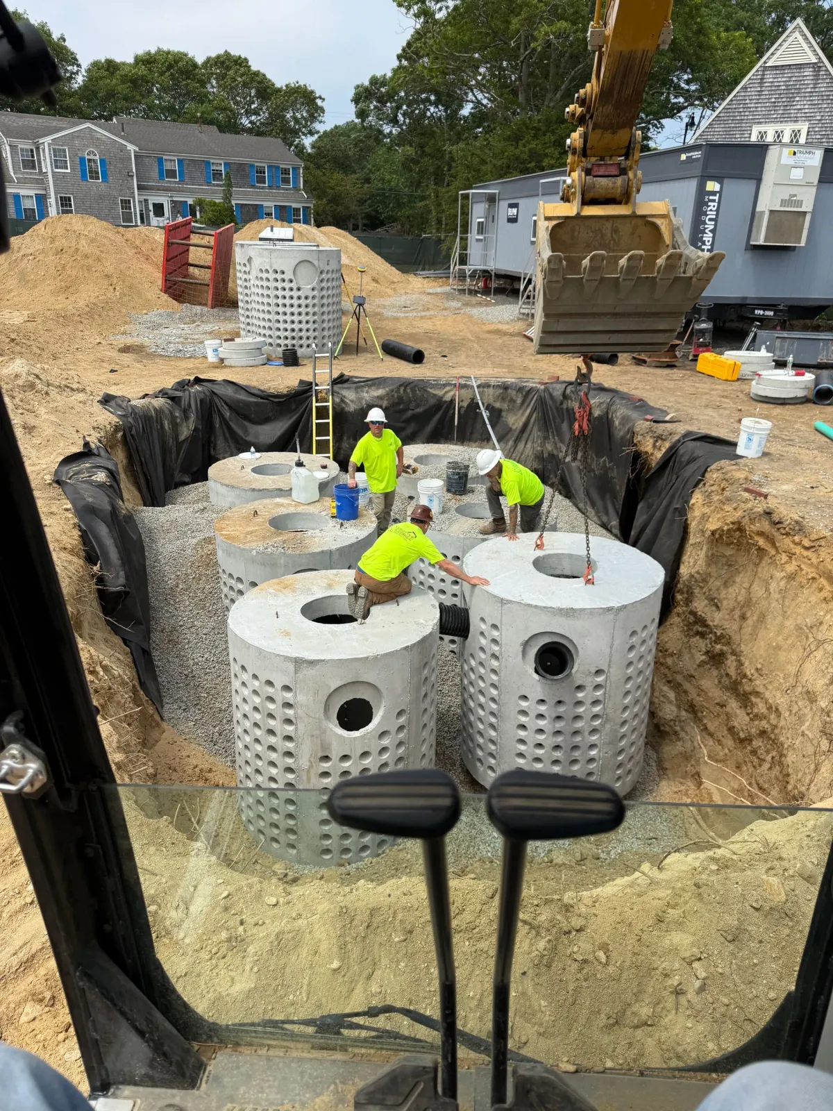 Construction site with workers installing large, cylindrical concrete tanks in an excavated pit. An excavator and buildings are in the background.