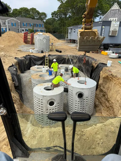 Construction site with workers installing large, cylindrical concrete tanks in an excavated pit. An excavator and buildings are in the background.