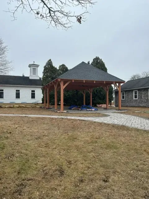 Wooden gazebo with dark roof, in a grassy yard near white buildings. Overcast day.
