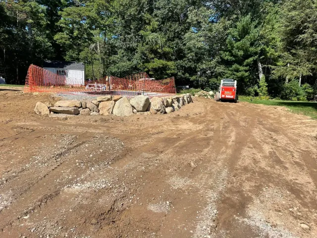 Construction site: Dirt road leads to a stone wall; orange fence and small bobcat sit in the background.