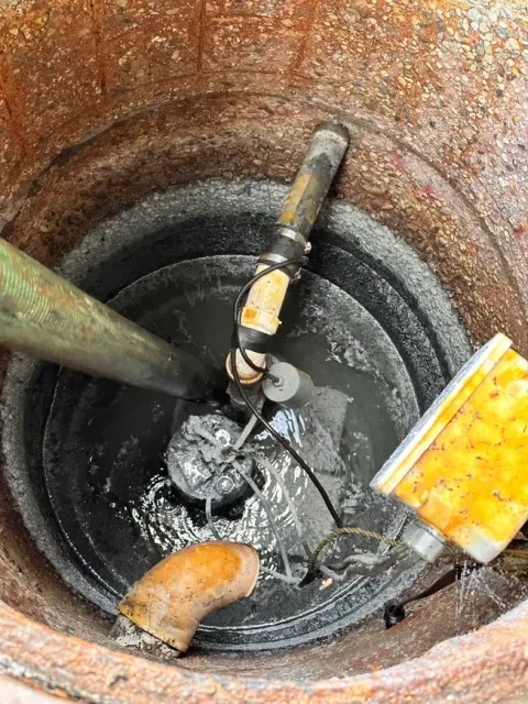Sump pump inside a dark, circular pit. Visible pipes, float switch, and debris in the water.