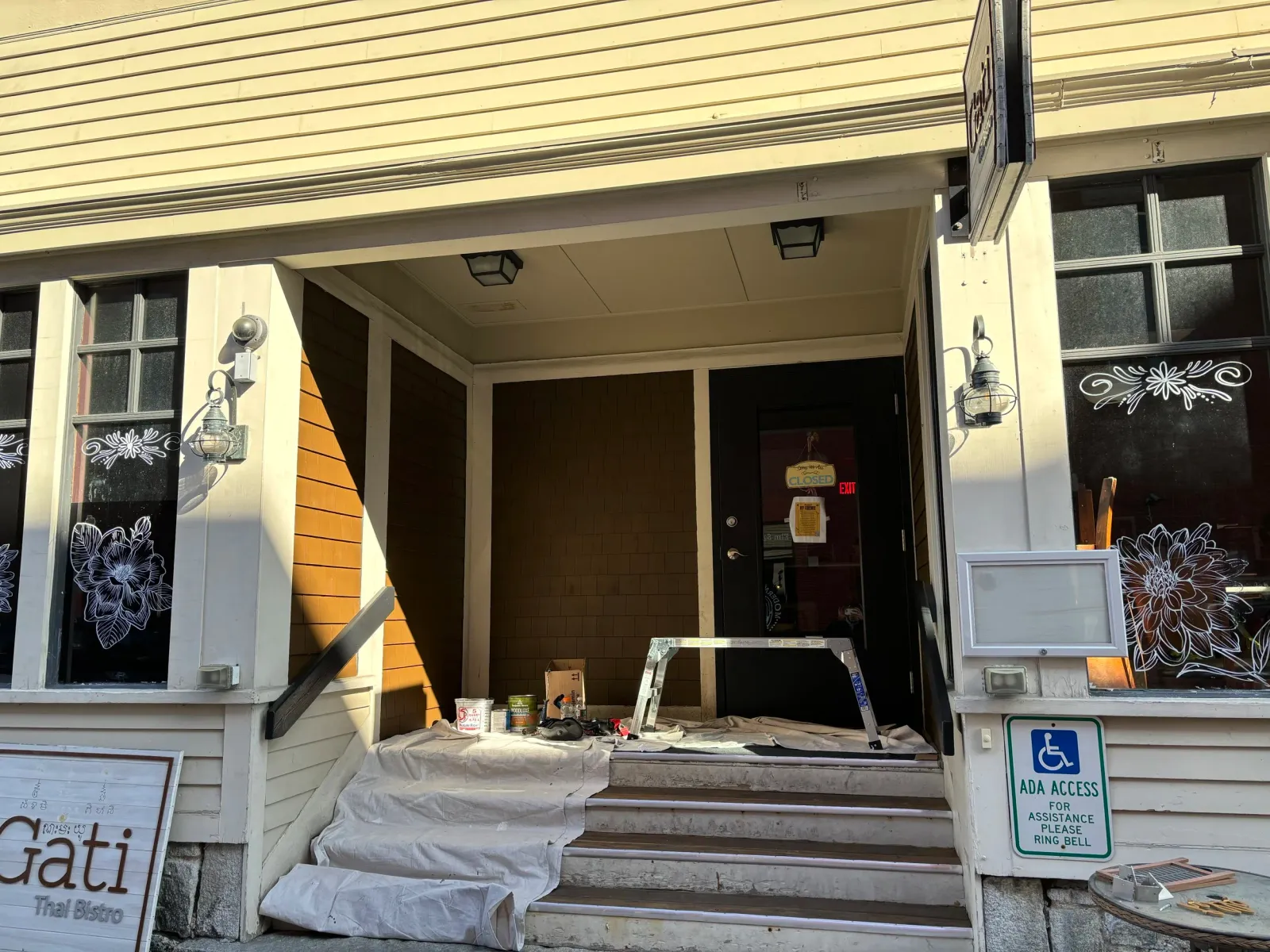 Exterior of a Thai bistro with entrance being painted, showing a closed door, paint supplies and a handicap access sign.