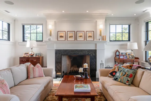 Living room with white walls, large windows, a fireplace, beige sofas with colorful throw pillows, and a wooden coffee table with candles and magazines.