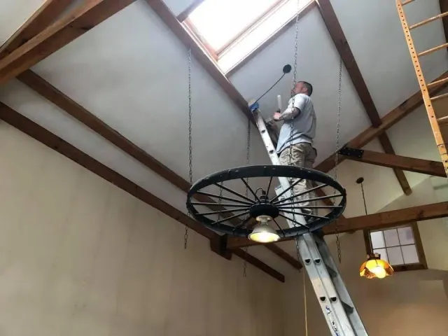 Worker on a suspended circular lift installing a skylight in a wood-beamed ceiling.