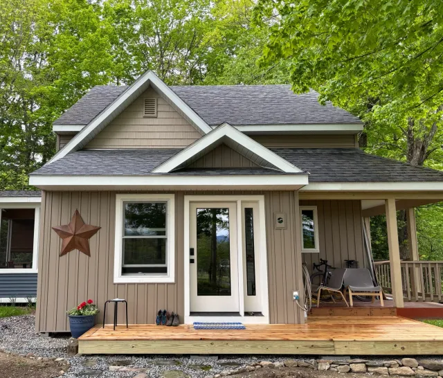 A small house with a front porch, beige siding, and a gabled roof surrounded by green trees, featuring a decorative metal star on the wall and outdoor chairs on the porch.