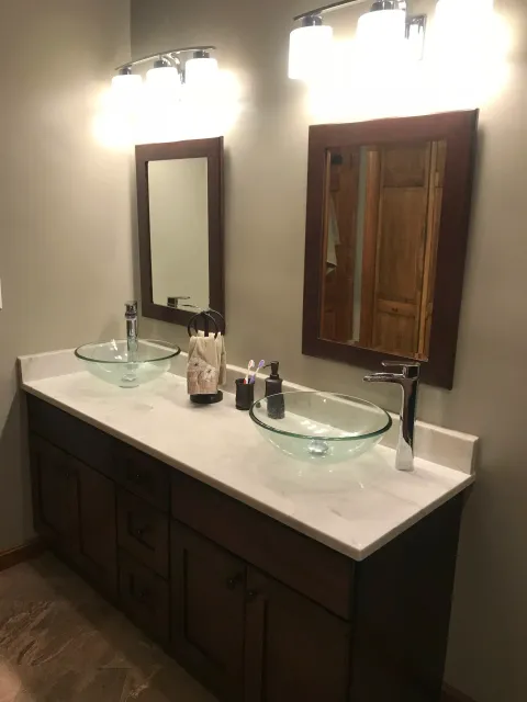 Bathroom double vanity with dark wooden cabinets, two round glass vessel sinks, modern chrome faucets, two rectangular mirrors with dark wooden frames, overhead light fixtures, and bathroom essentials like toothpaste and soap.