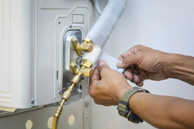 Hands connecting tubing to an outdoor air conditioning unit, showcasing copper fittings and white insulation.