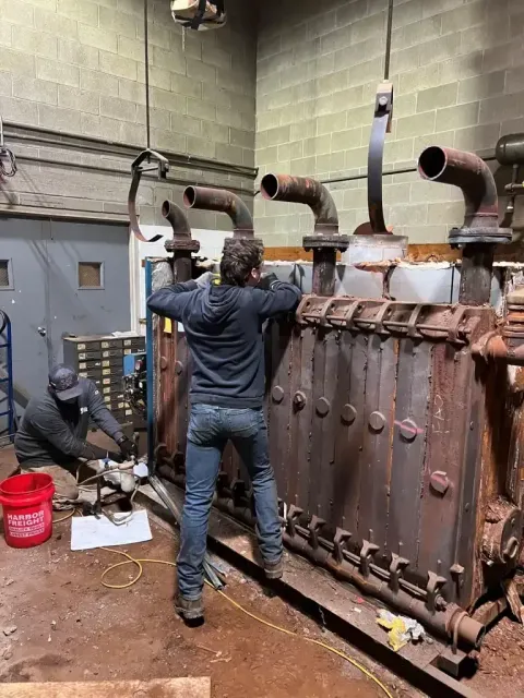 Two people work on a large, rusty industrial machine with vertical pipes in an indoor workshop.