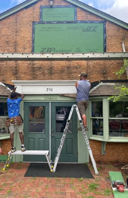 During: two crew members on ladders at a storefront entry, the front wall down to the sheathing with ZIP System panels in the upper gable