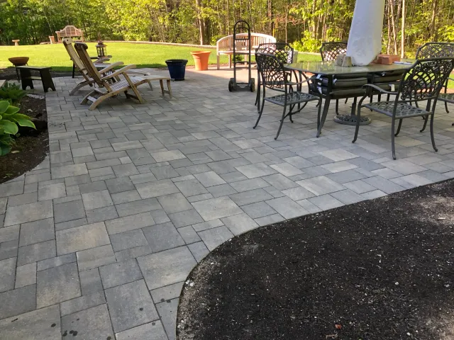 A backyard patio with stone tiles, outdoor chairs, a table, and potted plants, surrounded by green grass and trees in the background.