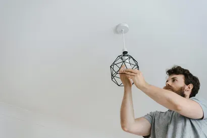 A person with a beard installing a light bulb into a black geometric pendant light fixture on a white ceiling.