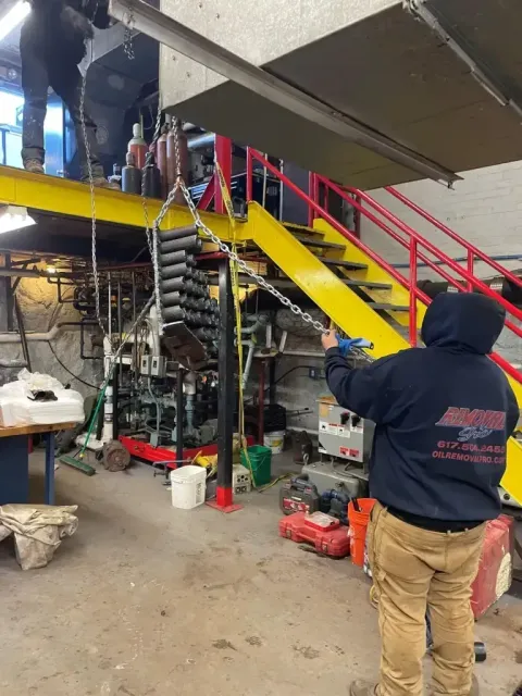 A person in a dark hoodie operates a chain hoist to lift a stack of circular mechanical parts inside a workshop.