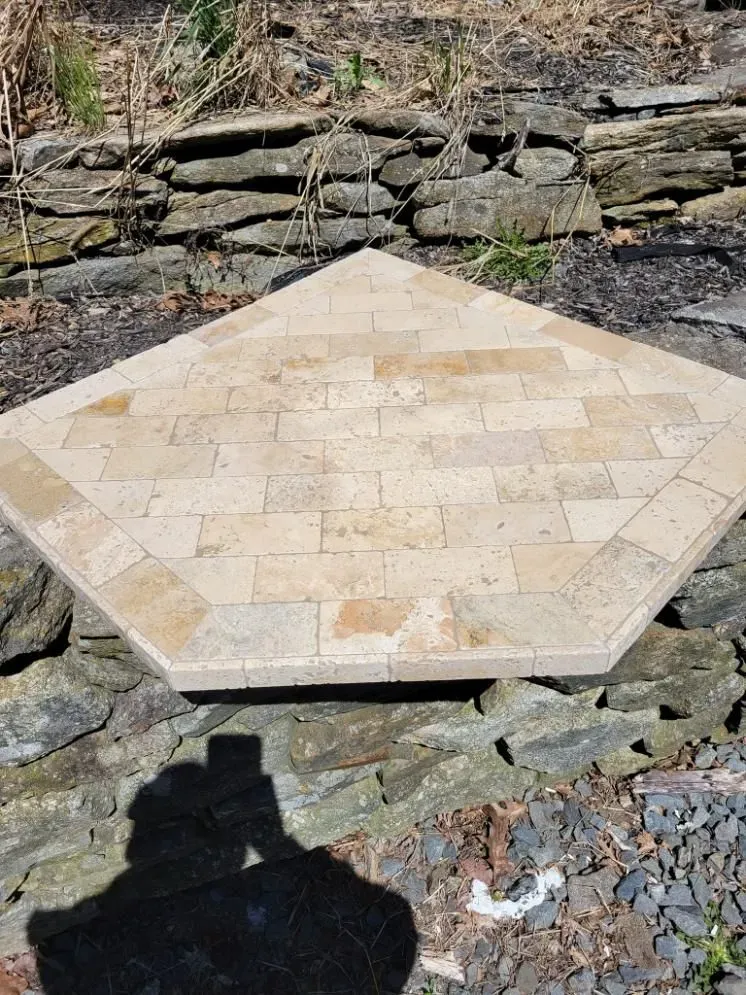 Beige hexagonal stone patio or table base on a rocky ground, with a stone wall behind.