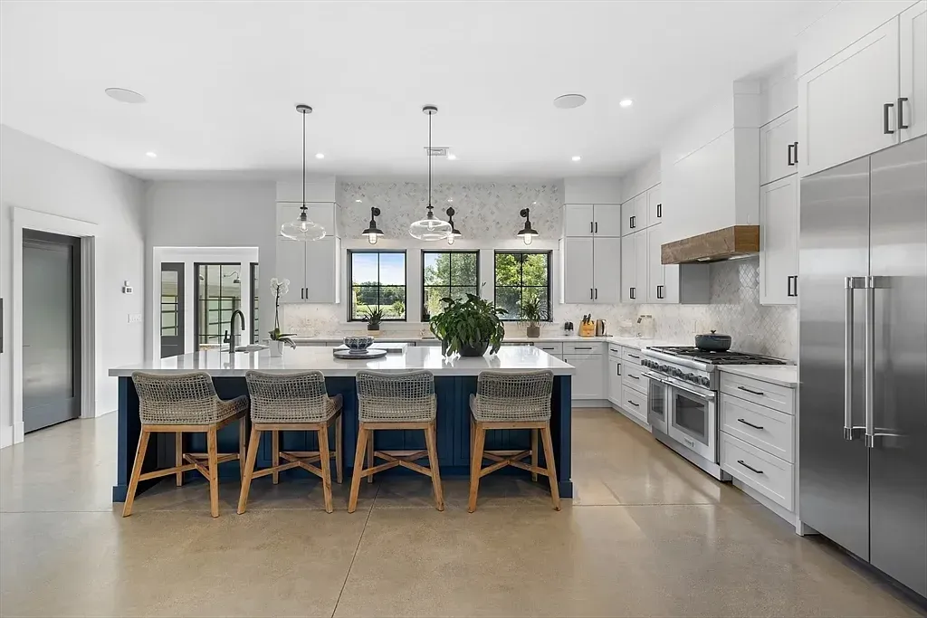 Modern kitchen with white cabinetry, stainless steel appliances, a large island with a dark blue base, five wicker chairs, pendant lighting, and a window with a view of trees outside.