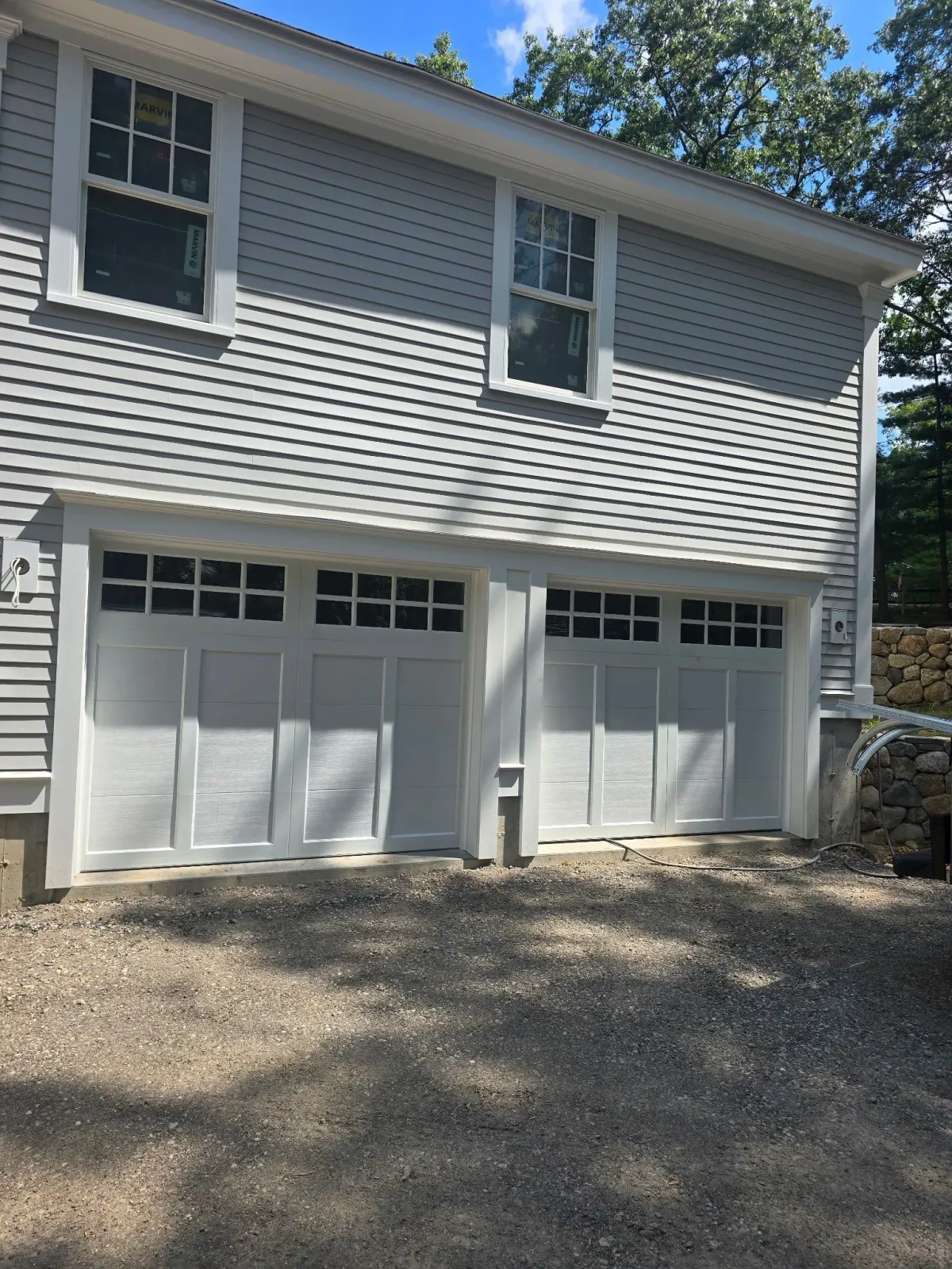 Two-story gray house with white trim and garage doors, windows, and a gravel driveway.