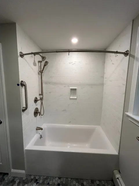 Bathroom with white bathtub, shower curtain rod, handheld shower, and grab bars in a tiled alcove.