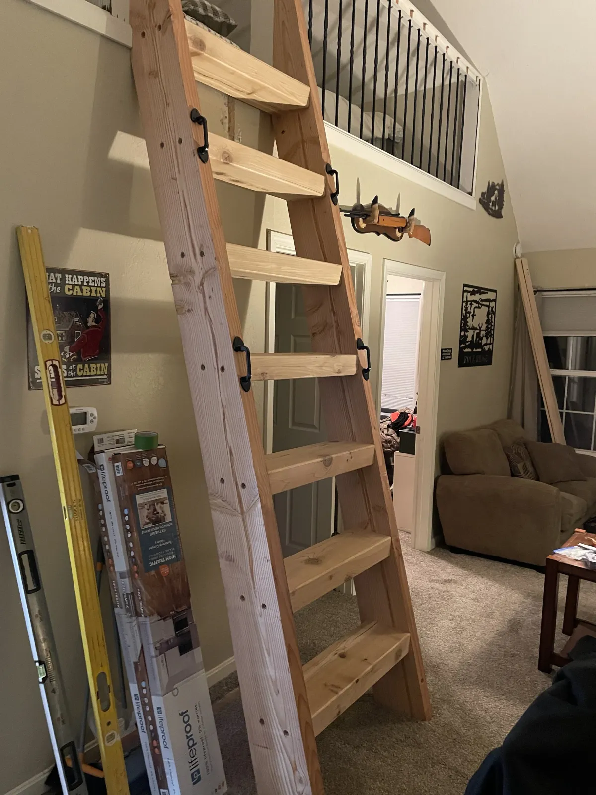 A wooden ladder leaning against a wall in a living room, with some tools and boxes nearby, and a second ladder resting against the wall. There is a staircase with a black railing above, and a foam kiddie pool with a toy gun attached to the wall. A br