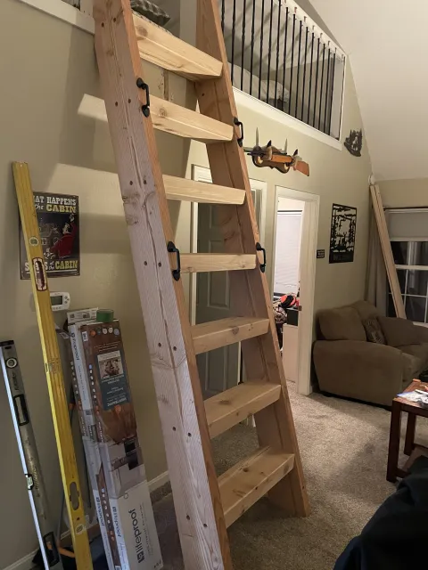 A wooden ladder leaning against a wall in a living room, with some tools and boxes nearby, and a second ladder resting against the wall. There is a staircase with a black railing above, and a foam kiddie pool with a toy gun attached to the wall. A br