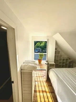 Bedroom with white walls, a bed, and a small balcony overlooking trees. Sunlight streams in.