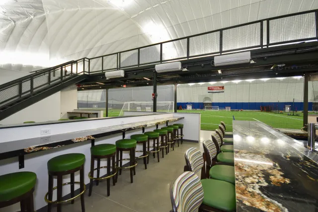 Indoor sports facility with a soccer field visible through large windows, a staircase with black railing leading to a second level, and a seating area with green barstools and a marble countertop.
