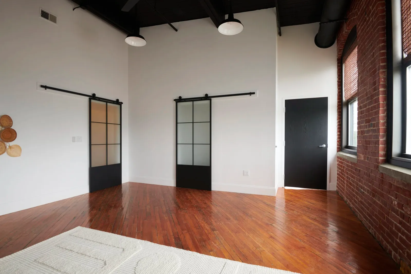 Empty room with white walls, wooden floor, exposed brick wall with two large windows, black door, and decorative wall hangings.