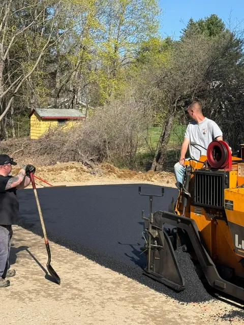 Two people work on a sunny day using a paving machine to lay fresh asphalt on a gravel driveway.