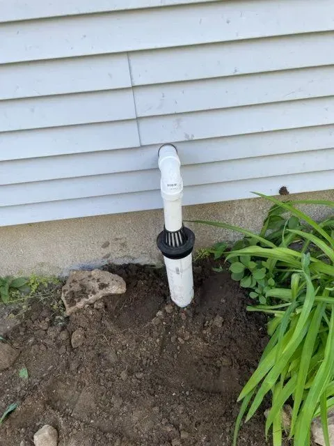 White PVC pipe with elbow fitting emerging from soil beside a house's vinyl siding and plants.