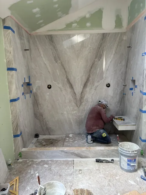A Benchmark tile setter fitting the bench inside a book-matched marble shower, mid-build