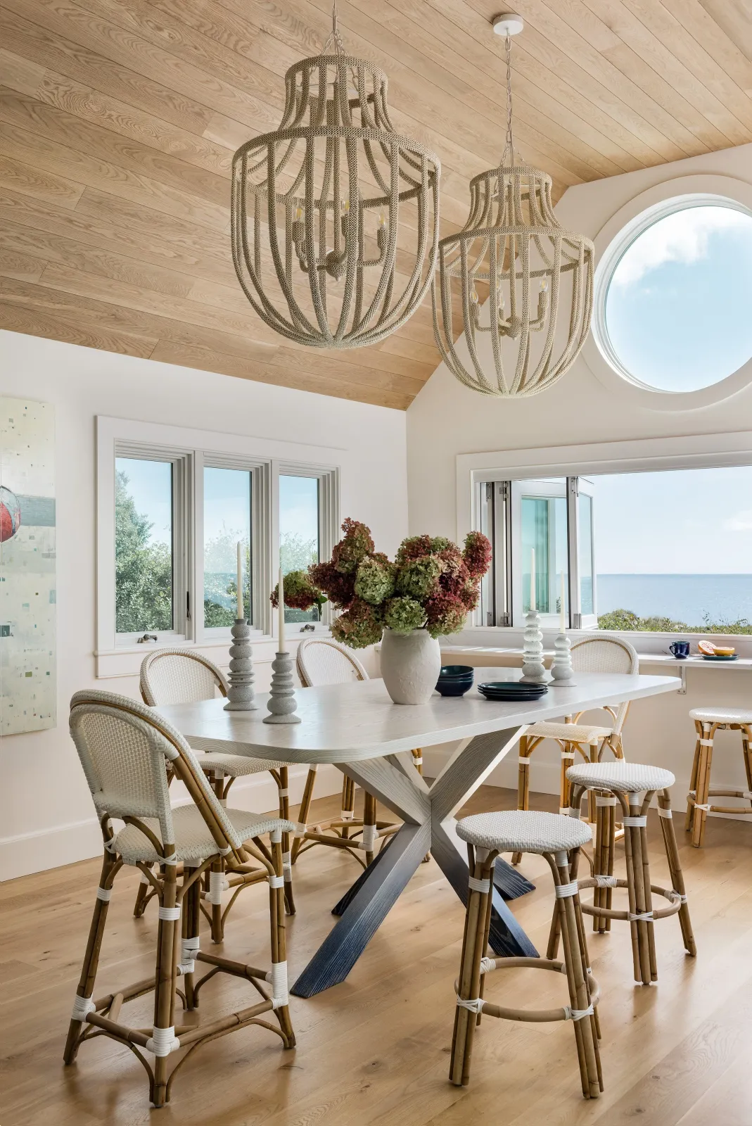 A bright dining room with a light wood ceiling, white walls, large windows showing a view of the ocean and sky. There's a white dining table with a vase of flowers, candlesticks, and black dishes, surrounded by white wicker and wood chairs. Two large