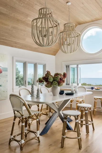 A bright dining room with a light wood ceiling, white walls, large windows showing a view of the ocean and sky. There's a white dining table with a vase of flowers, candlesticks, and black dishes, surrounded by white wicker and wood chairs. Two large