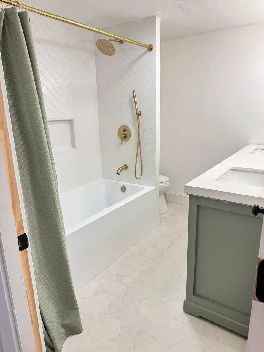 A modern bathroom with a white herringbone tiled tub, gold fixtures, a sage green vanity, and light patterned flooring.