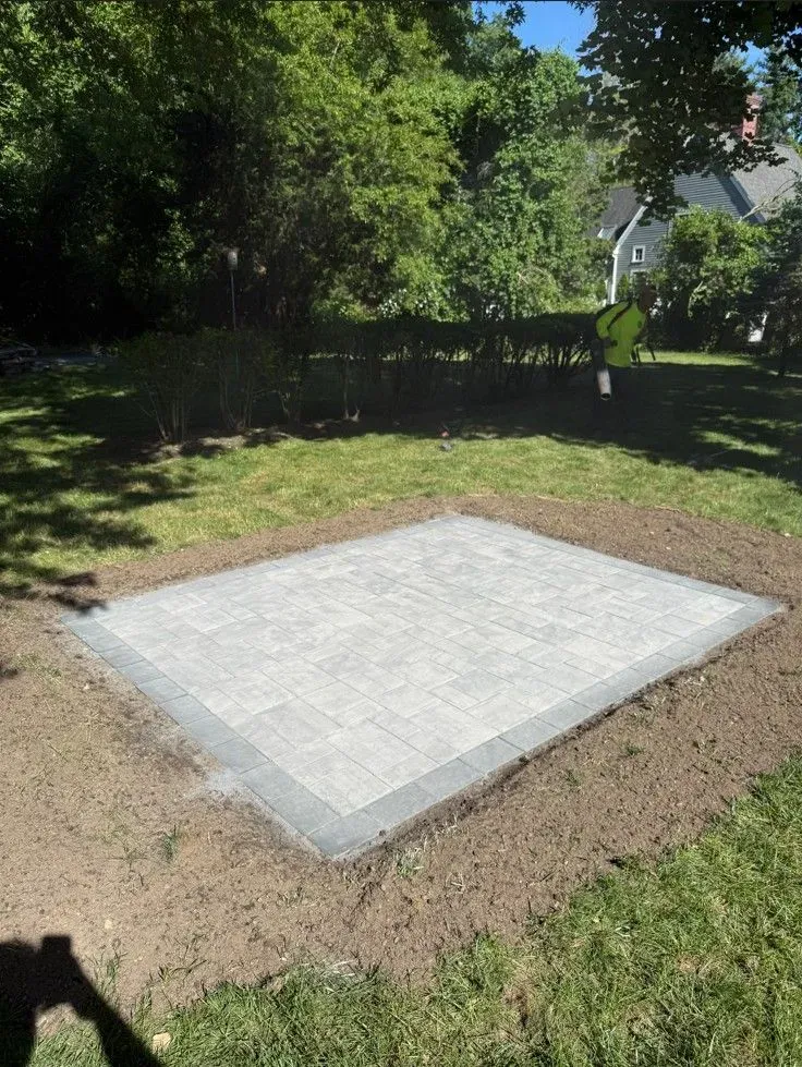Concrete slab in a grassy yard, bordered by dirt and trees in the background.