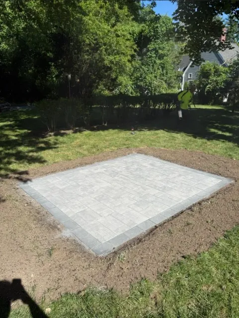 Concrete slab in a grassy yard, bordered by dirt and trees in the background.