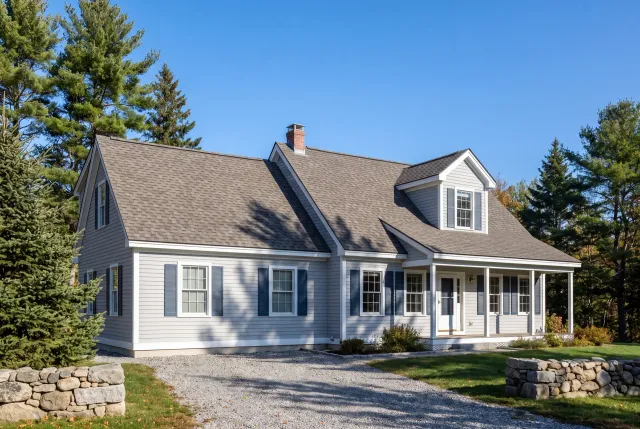 Roofing contractor: gray lap-sided cape with gray-brown arch shingles, gable dormer, blue shutters and farmer's porch