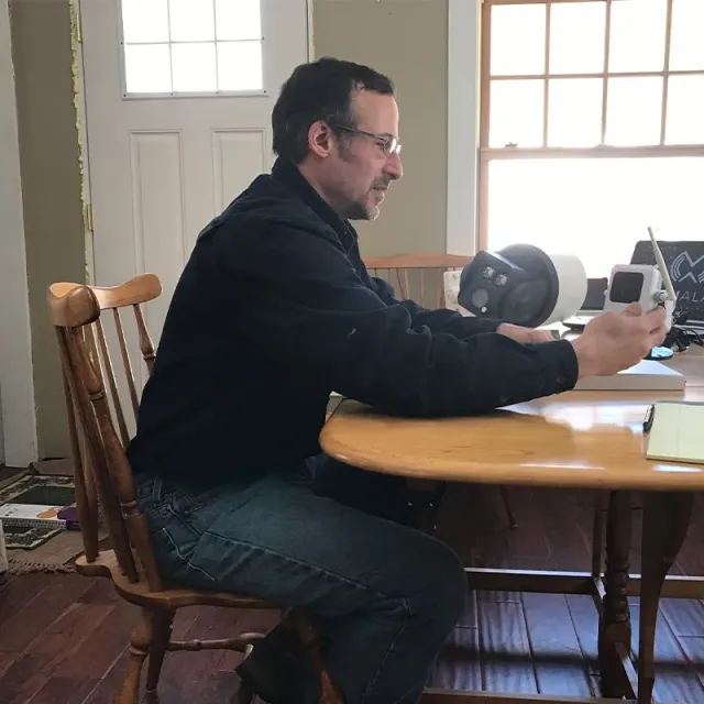 Man seated at table holding a small object, near a window and an opaque object.