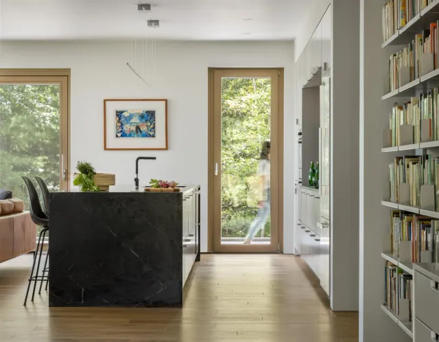 Modern kitchen with black marble island, wooden flooring, white cabinetry, and large windows showing green trees outside.