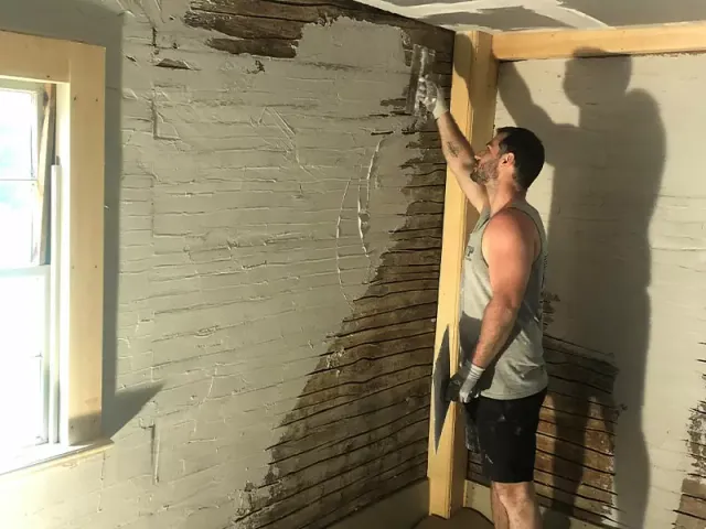 A person in a tank top and gloves applies plaster to a damaged wall inside a room. Sunlight streams through a nearby window.
