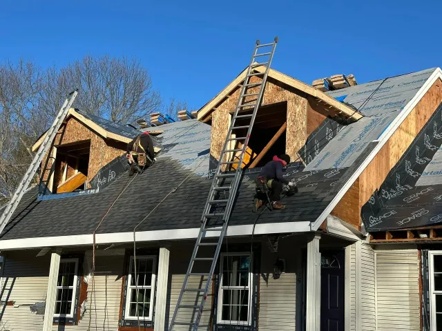 Workers repairing a house roof with ladders, exposed plywood, and dark shingles under a blue sky