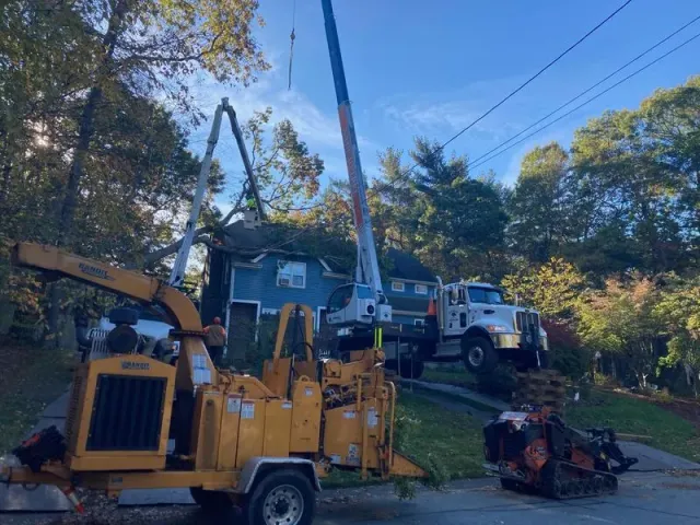 Yellow crane and utility truck working near a house on a wooded roadside with a small engine on the ground
