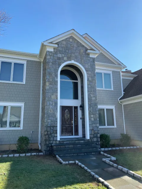 Front view of a modern house with stone and siding exterior, a black front door with decorative glass, and a small front porch with steps leading to a walkway. There are windows, bushes, and a lawn in the yard.