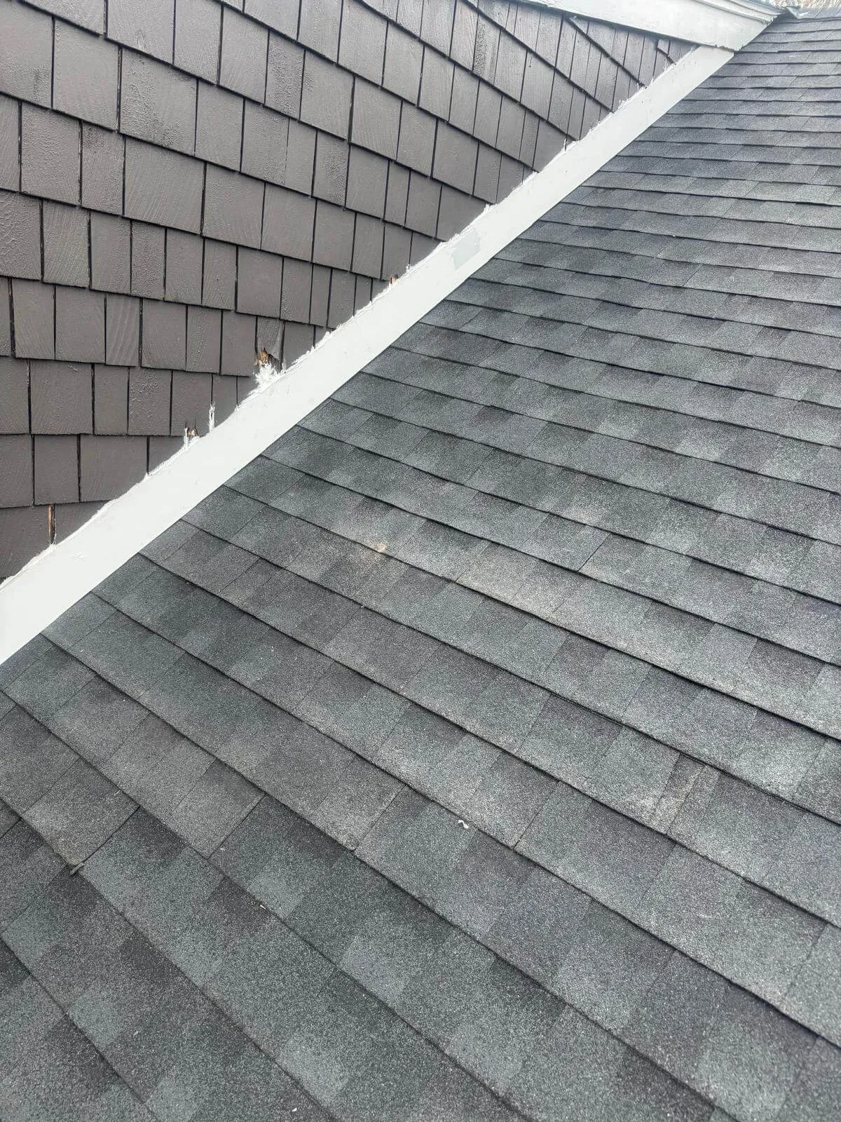 Close-up view of a residential roof showing gray asphalt shingles on the sloped surface and dark gray vertical siding shingles on the adjacent wall, with white flashing along the intersection.
