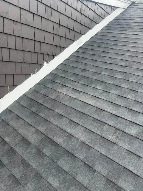 Close-up view of a residential roof showing gray asphalt shingles on the sloped surface and dark gray vertical siding shingles on the adjacent wall, with white flashing along the intersection.