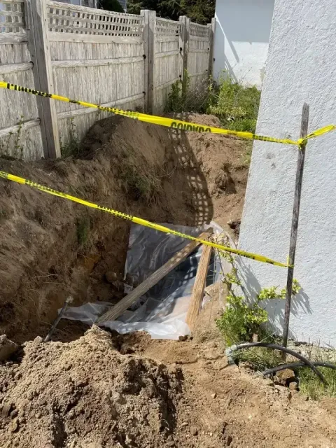 A deep trench with plastic lining sits next to a house wall, marked off by yellow caution tape.