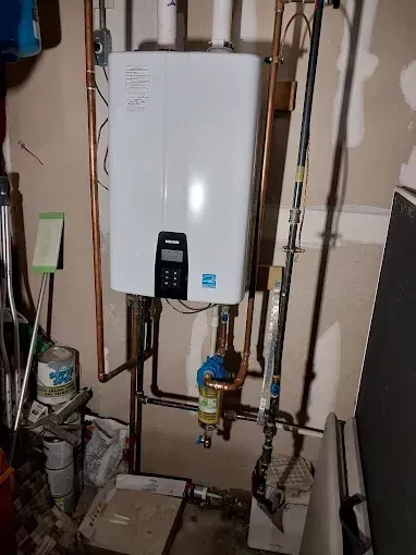 A wall-mounted white Navien tankless water heater with copper piping and a water filtration unit below it in a basement.