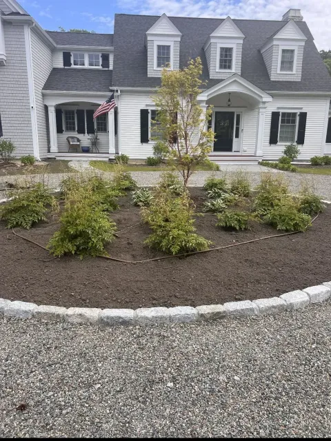 Gray house with dormer windows and a white porch behind a curved gravel driveway and landscaped shrubs