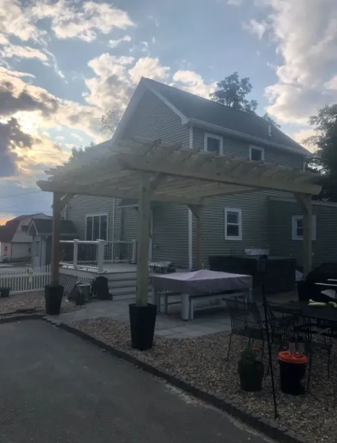 Wooden pergola over a patio with a covered table, in front of a green house.
