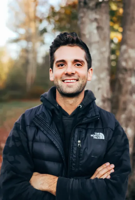 A man smiling outdoors wearing a black The North Face jacket, with blurred trees in the background.